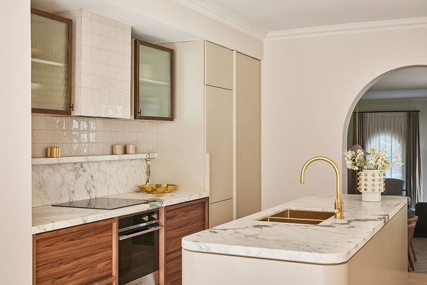 A refined, warm-toned kitchen with a large marble island, integrated sink and curved brass tap. The cabinetry combines cream-coloured tall units with dark timber-grain lower cabinets, while the rear wall features white tiled splashback, marble benchtops, open shelving and fluted-glass wall cabinets. An arched opening connects visually to a dining or living area beyond.