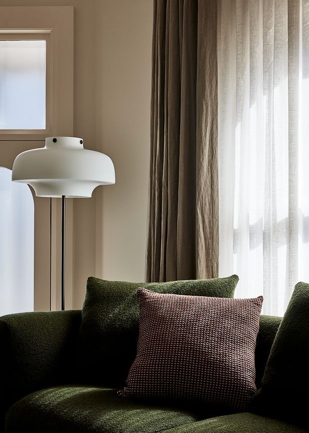 Close-up interior view of a dark olive-green upholstered sofa with textured green and burgundy cushions, positioned beside a large window with sheer white curtains and heavy taupe drapes. A sculptural white floor lamp and warm neutral walls create a refined, softly lit living area.