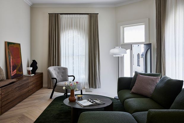A refined living room with a dark green upholstered sofa and armchair, round black coffee table, walnut-toned built-in sideboard, layered floor-to-ceiling curtains, and pale herringbone timber flooring. Classic cornicing and ceiling detailing combine with contemporary furniture and muted neutral finishes. Natural light enters through tall windows and a glazed door.