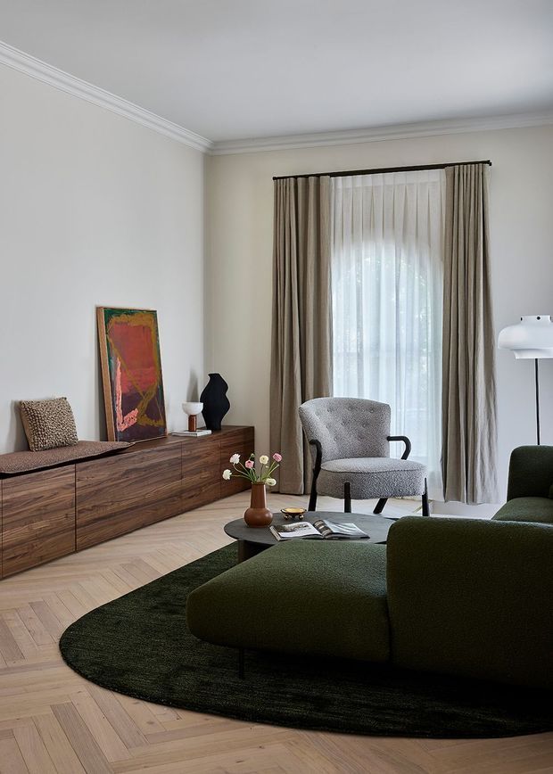 A refined living room with cream walls, decorative crown moulding, herringbone timber flooring, and full-height curtained glazing. A long dark timber sideboard with artwork and sculptural decor sits along the wall, while a textured grey armchair and deep olive-green upholstered seating surround a low black coffee table. A dark green oval rug anchors the seating area, creating a warm, sophisticated interior.