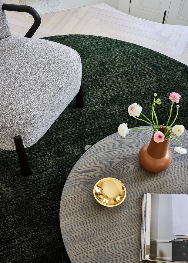 A styled living room vignette photographed from above, featuring a curved light-grey upholstered lounge chair with dark timber legs, a dark green textured rug, a round grey-toned timber coffee table, and a sculptural terracotta vase with flowers. Pale herringbone timber flooring and white paneled doors are visible in the background. The overall look is refined and contemporary with mid-century-inspired furniture forms.