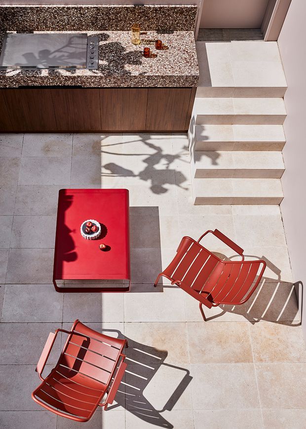 Sunlit rooftop or terrace entertaining area viewed from above, with a compact outdoor kitchen featuring a cooktop, mottled stone benchtop and dark timber cabinetry. Pale tiled flooring continues up a short flight of steps, while a red outdoor coffee table and two matching red metal lounge chairs create a bold seating arrangement. Strong angular shadows add a graphic, high-contrast quality.