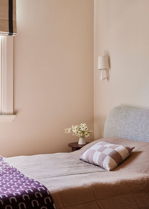 A softly styled bedroom with warm blush-beige walls, a textured upholstered bedhead, layered neutral bedding, and a mauve-and-cream checked cushion. A small dark timber bedside table holds a vase of white flowers, while a wall sconce and partially visible window provide additional detail. The overall atmosphere is calm, warm, and understated.