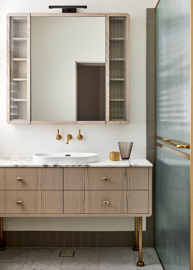 A refined contemporary bathroom vanity with a wall-mounted timber-look cabinet, white marble benchtop, and integrated white basin. Above it is a matching mirrored cabinet with fluted glass side compartments and a slim black light fixture. Brass wall-mounted tapware, handles, and slender vanity legs add warm metallic accents. The room features dark vertical wall tiles, pale marble-look floor tiles, and a textured fluted-glass door with brass hardware.