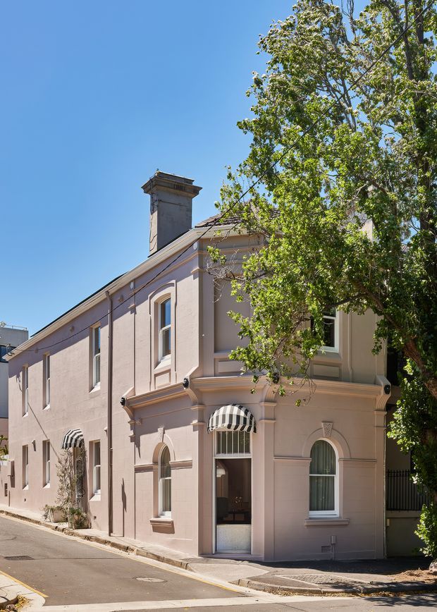 Sunlit street view of a narrow two-storey heritage terrace or townhouse with a pale pink rendered facade, ornate classical window surrounds, arched openings, striped awnings, a prominent chimney, and a mature street tree partially obscuring the front elevation. The building occupies a compact urban streetscape with a narrow road and footpath.