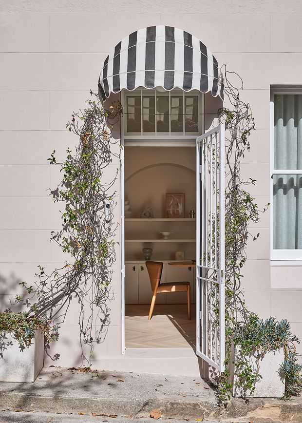 A charming residential facade with a pale rendered exterior and an open security gate leading into a compact study or home office. The entry is framed by climbing vines in planters, with a striped black-and-white scalloped awning above a multi-pane window. Inside, built-in arched shelving, a small desk, and a timber chair are visible. A neighbouring window with soft blue curtains appears at right.
