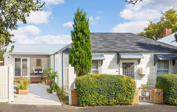 Front exterior view of a low-rise residential cottage-style home with pale weatherboard cladding, a dark hipped roof, decorative entry canopy and security door, brick garden walls, clipped hedges, and a paved path leading to a glazed side extension. The property has a traditional suburban character with neat landscaping and bright daylight.