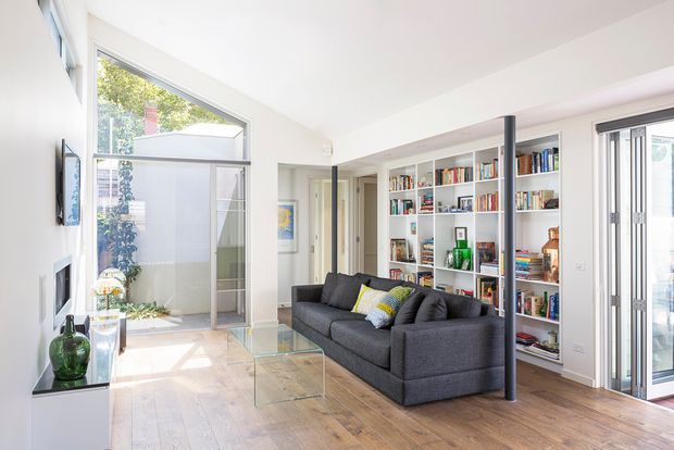 Bright contemporary living room with a dark charcoal sectional sofa, glass coffee table, extensive white built-in bookshelves, and warm timber flooring. Full-height glazing and a glazed courtyard connection bring abundant natural light into the open space, while a slim dark structural post adds contrast.