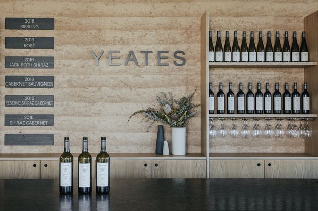 A refined wine bar or tasting room with a textured beige stone feature wall, prominent YEATES lettering, built-in timber shelving displaying wine bottles and hanging glasses, and a dark stone counter. Three wine bottles sit on the foreground counter, while a small floral arrangement and award plaques add detail.