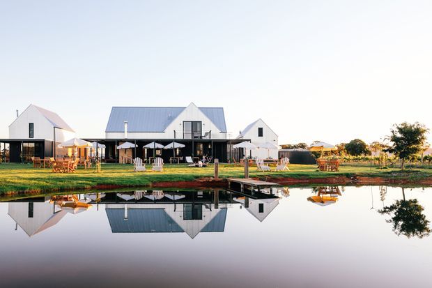 A wide exterior view of a refined rural or hospitality-style property beside a calm reflective pond. Several white gabled buildings with steep pitched roofs and dark-framed glazing are arranged around a landscaped lawn, with outdoor seating, umbrellas, and dining areas along the water. The scene has a bright, serene, polished appearance and a strong symmetrical reflection.