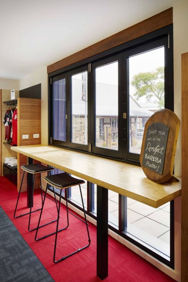A compact, contemporary café or hospitality interior with a long pale timber counter positioned in front of large black-framed windows. Several tall black bar stools line the counter, while a bold red carpet defines the seating zone. Timber wall panels, a dark ceiling-mounted shelf, and a chalkboard sign create a warm, informal workspace or bar-like setting. The windows overlook a courtyard and neighbouring building.