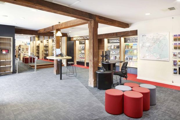 Interior view of a visitor information centre or tourism office, with exposed heavy timber posts and beams, carpeted flooring, reception and service desks, brochure shelving, information signage, a wall map, and clustered round upholstered stools. The open-plan commercial space has a bright, functional fit-out with red accent carpeting and seating.