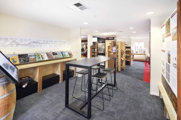 A bright, contemporary commercial showroom or customer display area with grey carpet flooring, white walls and ceiling, recessed downlights, and a central black-framed high table with bar stools. Product and information displays line the walls, with shelving and retail-style fixtures extending toward the rear. A red carpet runner provides a strong accent along the right side.