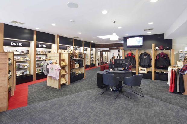 A bright, modern retail interior with a central checkout or service desk, display shelving, clothing racks, branded product areas, dark feature walls, timber-framed joinery, and grey patterned carpet. Red flooring accents define circulation zones, while recessed ceiling lights and a suspended pendant provide even illumination.