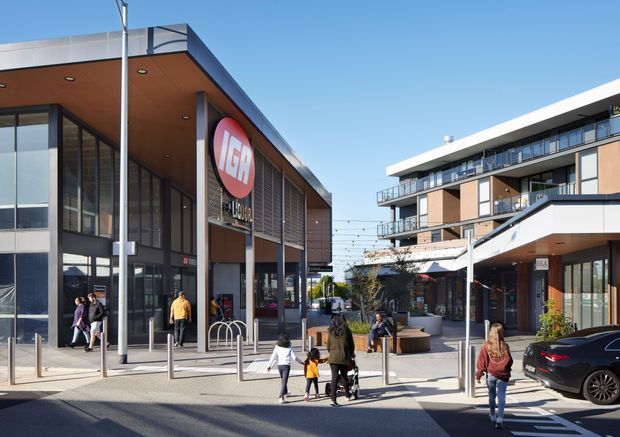 A contemporary mixed-use urban streetscape with a modern retail building on the left and multi-storey apartment or commercial buildings on the right. The scene includes glazed storefronts, timber-toned soffits, dark metal framing, balconies, paved pedestrian areas, bollards, outdoor seating, signage, and parked vehicles. Bright clear daylight emphasizes the active public plaza and pedestrian connection.