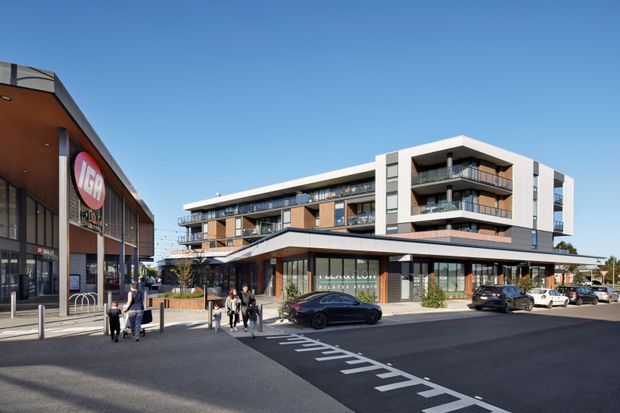 Exterior streetscape view of a contemporary mixed-use development with a multi-storey apartment building above ground-floor retail and commercial tenancies. The complex has white rendered and dark metal facade elements, projecting balconies, extensive glazing, and a flat roof. A large retail building, paved forecourt, driveway markings, pedestrians, bicycles, and parked vehicles are visible.