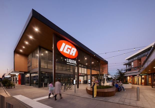 Dusk exterior view of a contemporary IGA supermarket and liquor store in a pedestrianised commercial plaza. The building features a dramatic angular cantilevered roof, dark metal framing, extensive floor-to-ceiling glazing, warm timber-toned soffits, and illuminated signage. Adjacent retail buildings, paved public space, outdoor seating, planter beds, bicycles, and festoon lighting are visible.