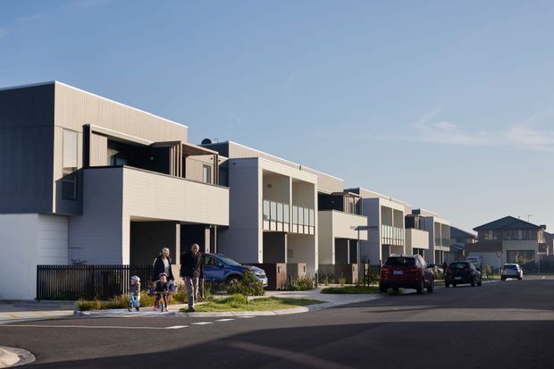 Street-level exterior view of a contemporary row of attached multi-storey townhouses. The development features crisp geometric massing, flat rooflines, pale fibre-cement or rendered facade panels, dark recessed balcony areas, and integrated car parking along a newly paved suburban street. A detached pitched-roof house is visible in the background.