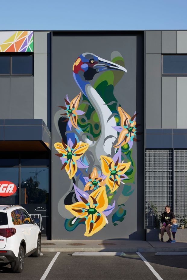Exterior view of a contemporary commercial building facade featuring a large, colourful bird-and-floral mural set within a tall dark recessed panel. The surrounding facade uses grey panel cladding, dark window frames, glazed storefront elements, and a gridded metal screen. A marked parking area and people are visible in front of the building.