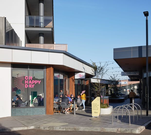 A contemporary mixed-use commercial precinct viewed from an outdoor pedestrian walkway. The scene features low-rise retail tenancies with modern white and dark panelled facades, timber-look vertical cladding, balconies with glass balustrades, outdoor café seating, bicycle racks, signage, and landscaped planter beds under a clear sky.