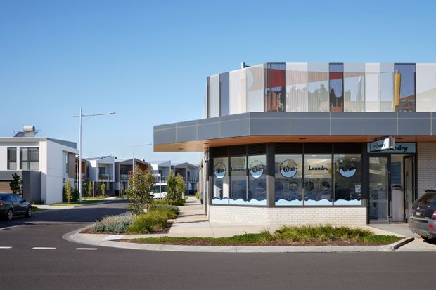 A contemporary low-rise commercial building photographed from a paved street, with a ground-floor laundromat and a projecting upper level enclosed by translucent and clear glazing. The facade combines dark horizontal panels, pale brickwork, white framing, and warm timber-toned soffit detailing. Modern residential-style buildings, landscaped verges, parked cars, and a clear blue sky surround the site.