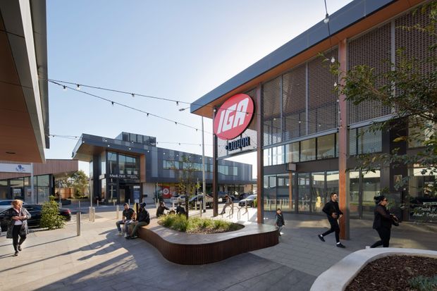 A contemporary open-air shopping centre courtyard with multi-storey commercial buildings, large glazed shopfronts, dark metal framing, and a prominent liquor retail sign. The paved plaza includes curved built-in planters, seating, pedestrian circulation, and overhead festoon lighting. The scene is bright and urban, with visible retail tenancies and landscaping.
