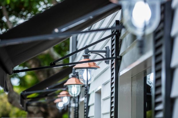 Close-up exterior architectural detail showing a row of wall-mounted lantern lights beneath dark awnings along a light-coloured weatherboard-clad facade. The image focuses on the facade, repetitive brackets, glazing, and outdoor lighting, with greenery softly blurred in the background.