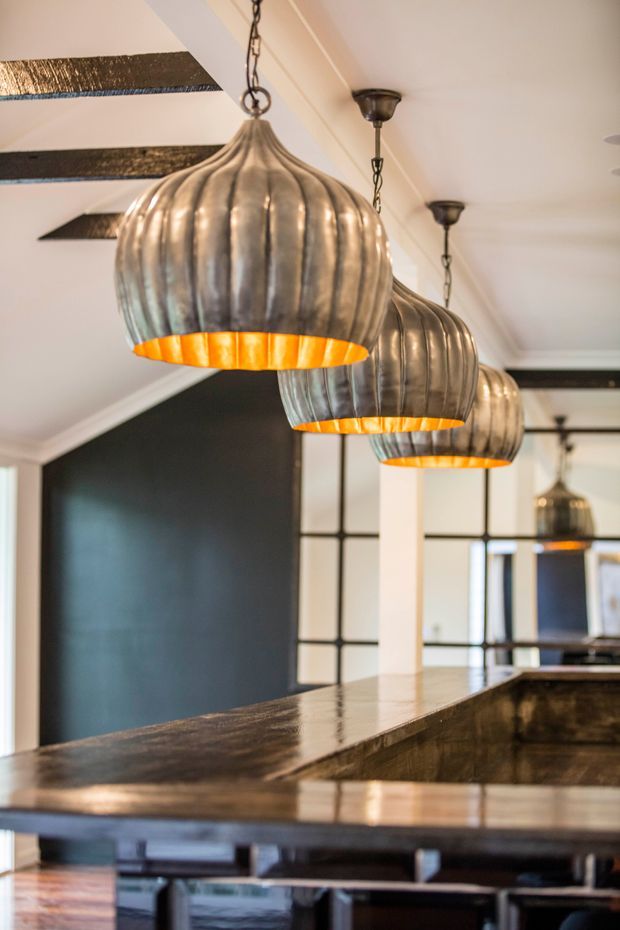 Close-up interior view of a sophisticated kitchen or bar island, featuring a dark polished stone benchtop, a deep dark feature wall, black-framed internal glazing, exposed dark timber ceiling beams, and a row of sculptural ribbed metal pendant lights with warm orange interiors. The space has a refined, dramatic, industrial-inspired character.