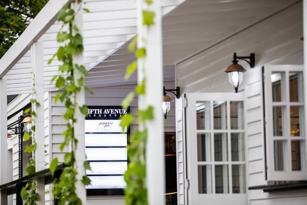 Close-up exterior view of a white weatherboard-clad building with a covered entry or verandah, multi-pane glazed doors and windows, black wall-mounted lanterns, and climbing greenery around a white timber frame. A partially visible sign identifies the venue as Fifth Avenue.