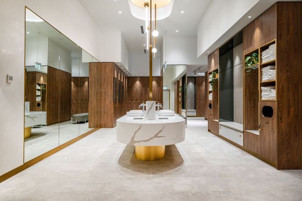 A spacious, luxurious bathroom with a central sculptural double vanity, integrated basins and a gold cylindrical pedestal. Full-height mirrors line the left wall, while dark timber wall cabinetry, open towel shelving, black textured panels and warm integrated lighting define the right side. The room features a light polished stone or concrete-look floor and white walls, with a large walk-in shower visible in the mirror reflection.