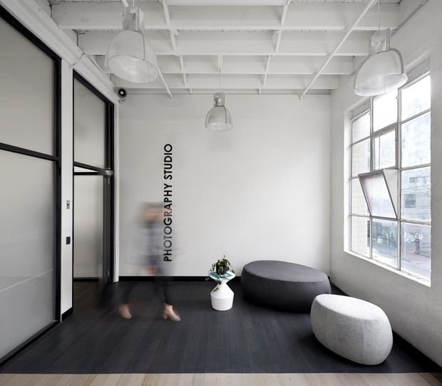 Bright minimalist studio interior with white walls, exposed white-painted ceiling beams, large industrial-style steel-framed windows, black-framed glazed partitions, and a dark timber floor. Rounded charcoal and light-grey upholstered lounge ottomans sit beside a small white side table and plant. A blurred person and vertical photography-studio lettering are visible, suggesting a creative photography workspace.