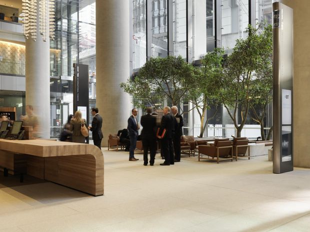 A spacious contemporary commercial lobby or atrium with a large timber reception desk, polished light flooring, tall cylindrical structural columns, extensive floor-to-ceiling glazing, indoor trees, lounge seating, and sculptural pendant lighting. Several people are gathered near the seating area, and a multi-level glazed interior is visible in the background.