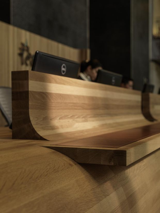 Close-up interior view of a sophisticated reception desk or service counter, constructed with prominent warm-toned timber joinery and a layered, sculptural curved edge. Computer monitors and staff are visible behind the desk, with dark wall panels and vertical timber detailing forming the backdrop. The image emphasizes craftsmanship, grain variation, and refined commercial fit-out design.