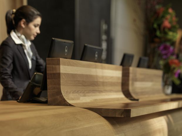 A close-up view of a reception desk in a commercial lobby, featuring sculptural curved timber joinery with layered wood grain and integrated computer workstations. A receptionist is visible behind the desk, with floral arrangements in the background.