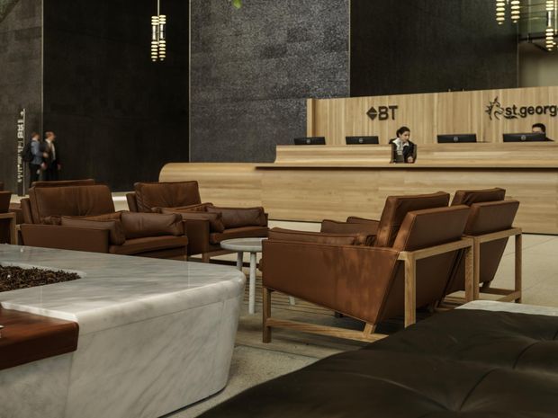 A sophisticated hotel or commercial reception lobby with a long light-wood reception desk, dark textured wall panels, marble-clad low seating or counter in the foreground, and multiple brown leather lounge chairs arranged around small tables. Reception staff and corporate logos are visible behind the desk. The space has a refined, contemporary hospitality design with warm timber and leather contrasted against charcoal surfaces and polished metal lighting.