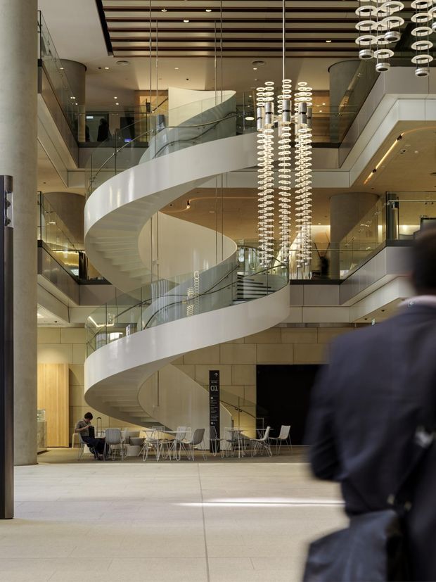 A grand multi-level commercial atrium with a prominent white sculptural spiral staircase, transparent glass balustrades, exposed cylindrical columns, and extensive mezzanine circulation. Numerous suspended ring pendant lights form a dramatic vertical installation beneath a slatted timber ceiling. Café-style tables and chairs occupy the ground level, while people and office-like activity are visible around the atrium.