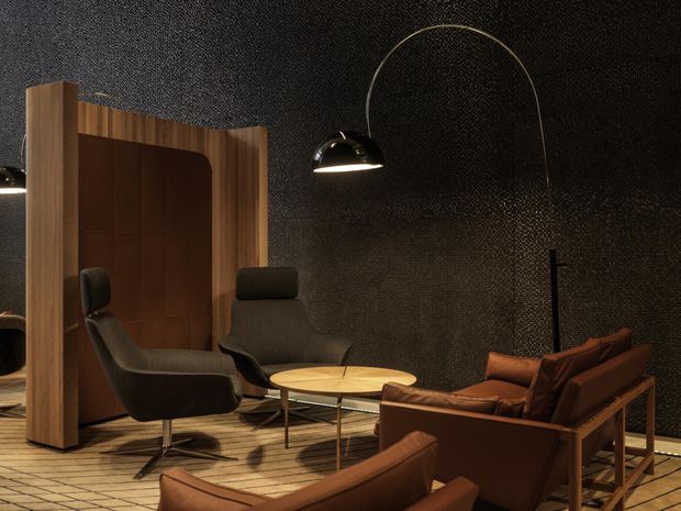 A dark, refined lounge or reception area featuring deep brown leather lounge chairs and sofas around a low round timber coffee table. The space has a dramatic black textured wall and ceiling, warm timber flooring, a sculptural timber room divider with curved detailing, and oversized arched black pendant lighting. The overall composition is intimate, luxurious, and strongly mid-century inspired.