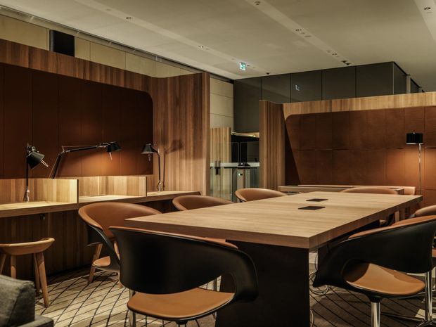 A refined contemporary meeting or dining room with a large light-wood rectangular table surrounded by dark sculptural chairs. Rich walnut-toned wall panelling, integrated shelving and cabinetry, warm brown upholstered feature panels, black task lamps, and a patterned carpet create a warm, sophisticated workspace atmosphere. A glazed or open partition and accessibility signage are visible in the background.