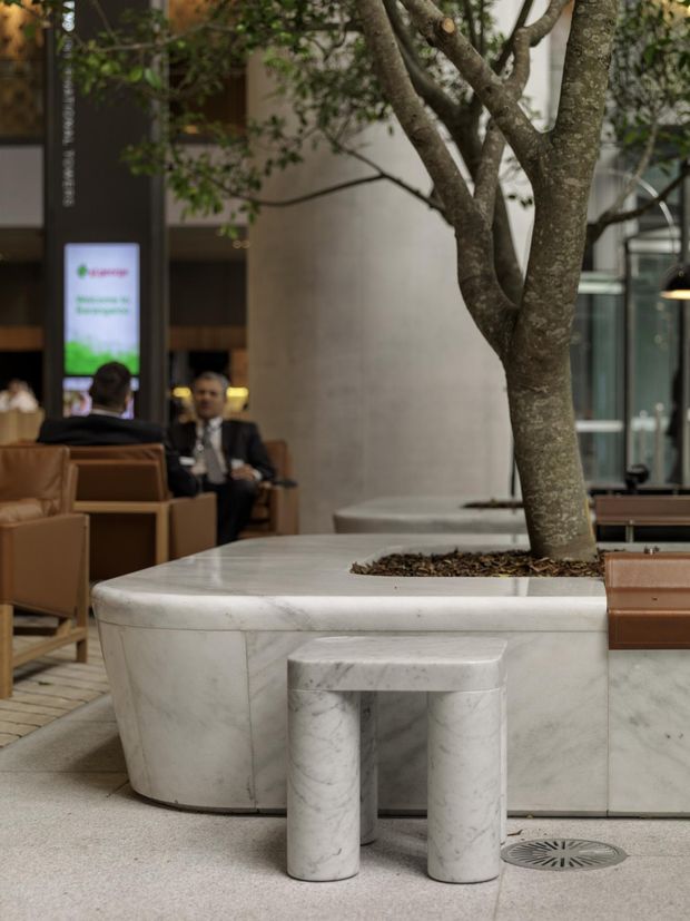 A sophisticated commercial interior lobby or atrium featuring a large integrated white marble planter and curved marble seating structure surrounding an established indoor tree. Warm tan upholstered lounge chairs, dark architectural columns, glass walls, and subdued lighting create a refined hospitality or office reception atmosphere. People and a digital sign are visible in the background.