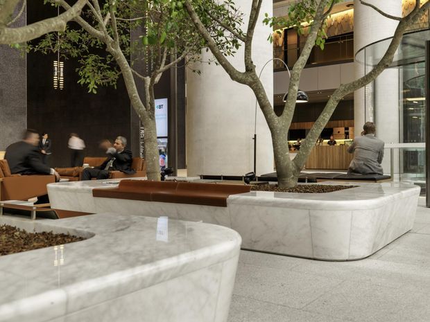 A spacious contemporary commercial interior lobby or atrium with integrated white marble or stone planters containing mature indoor trees. Curved built-in planter seating with brown leather or upholstered bench tops surrounds the planting beds. People sit and move through the space, with a reception or café-like timber counter and multi-level interior visible in the background. The space has polished light terrazzo or stone flooring, warm architectural lighting, tall glazing, and a refined hospitality or workplace atmosphere.