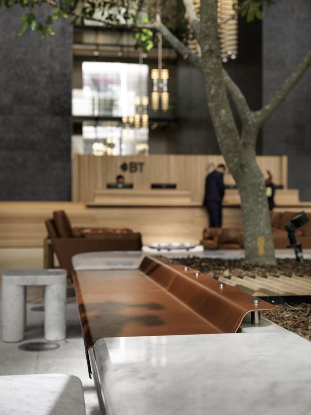 A sophisticated commercial lobby or reception area with a large indoor tree integrated into a landscaped planter. The foreground features sculptural built-in seating and tables combining pale stone or concrete with warm copper-toned metal and leather-like upholstery. A timber reception desk, lounge seating, dark textured walls, suspended pendant lighting, and a high glazed volume create a refined hospitality or workplace entrance.