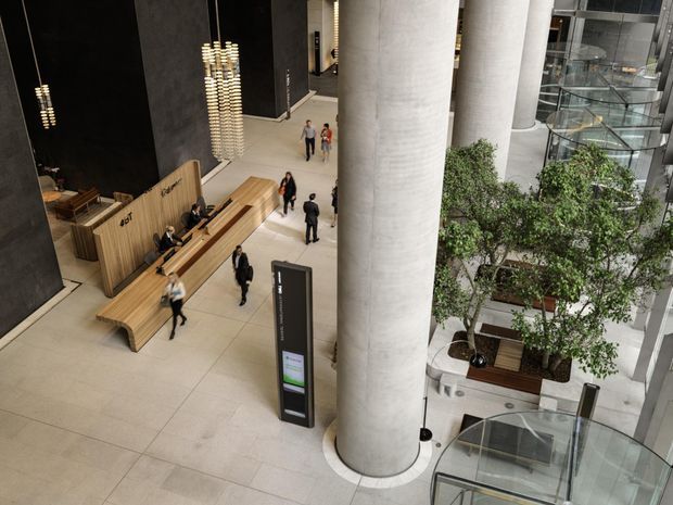 A large, modern multi-storey commercial lobby or atrium viewed from an elevated position. The space features expansive pale stone or concrete-look tiled flooring, monumental round concrete columns, black wall panels, a timber reception desk and integrated timber bench seating, sculptural hanging lights, indoor trees, glass balustrades, and a visible escalator or glass lift structure. People are dispersed through the spacious public interior.