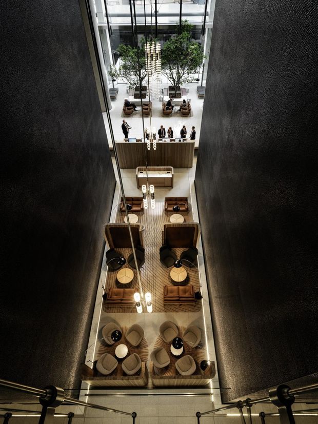 A dramatic high-angle view into a tall, narrow atrium or lobby of a contemporary commercial interior. Dark textured walls frame a multi-level hospitality or reception space with timber flooring, a large reception desk, lounge seating, potted indoor trees, and multiple hanging pendant light clusters. The space appears refined and hotel-like, with visible glass balustrades and a strong vertical void.