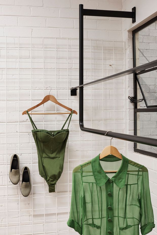 A close-up of a fashion retail or wardrobe display against a white-painted brick wall. Black metal hanging rails project from the wall and support wooden clothes hangers displaying green garments, while a pair of shoes is mounted on the wall. A black-framed industrial-style window is visible at right, reinforcing the utilitarian retail or studio setting.