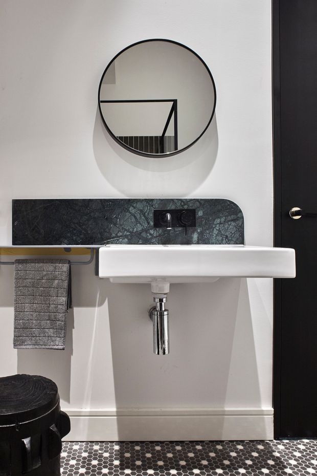 A compact, refined bathroom vanity area with a wall-mounted white ceramic basin, polished chrome plumbing, round wall mirror, dark marble-look splashback, black door, and patterned monochrome mosaic floor tiles. A grey hand towel hangs beside the basin, and a dark round stool or bin is partially visible in the foreground.