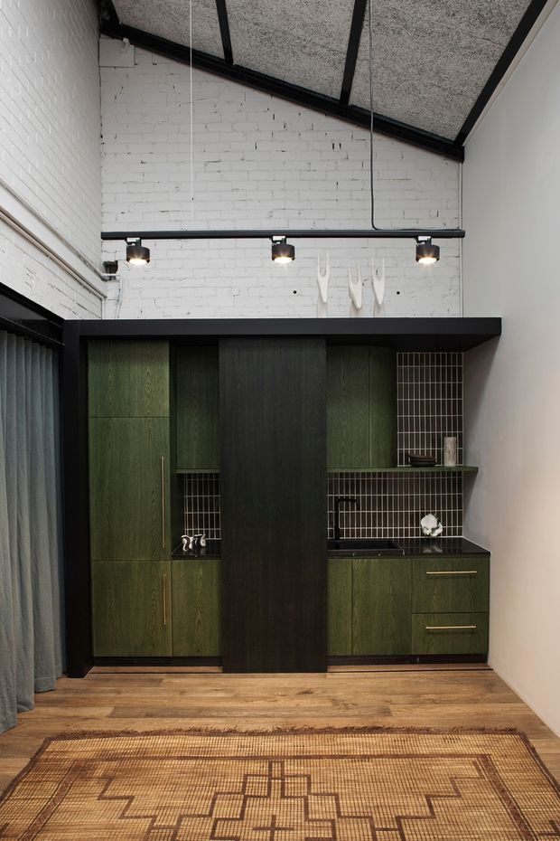 A compact, moody built-in kitchenette or bar-style joinery installation set against exposed white-painted brick walls. The cabinetry is dark green woodgrain with a tall black central panel, black benchtops, and small dark tiled splashbacks. Brass-toned handles, open shelving, track lighting, exposed black ceiling beams, timber flooring, and a geometric woven rug create an industrial yet refined interior.