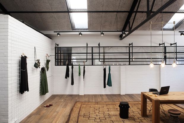 A bright industrial-style clothing studio or showroom with white-painted brick walls, exposed dark ceiling structure, skylights, track lighting, black-framed internal glazing and balustrades, and pendant lights. Garments hang along the walls and from rails, while a timber worktable with a laptop occupies the foreground. The space has warm timber flooring and a patterned rug.