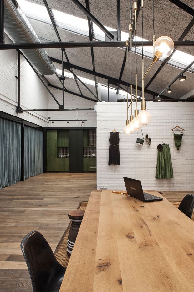 An open-plan fashion showroom or studio with an industrial warehouse character. The space features exposed black steel roof framing, acoustic ceiling panels, skylights, visible HVAC ductwork, white painted brick walls, timber flooring, dark curtains, green and black built-in cabinetry, and a long solid timber communal worktable with black chairs. Pendant globe lights hang above the table, while clothing is displayed on the white brick partition wall.
