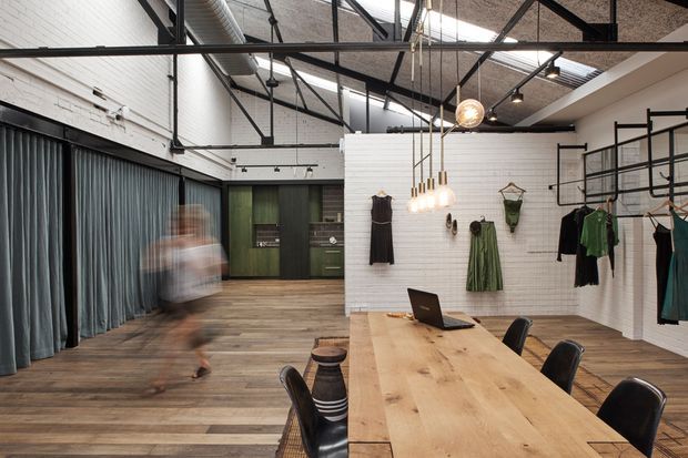 An industrial-style retail showroom or studio with exposed black steel roof trusses, ductwork, track lighting, and a high textured ceiling. White-painted brick walls divide the open space, with dark curtains, green cabinetry, timber plank flooring, and wall-mounted clothing displays. A large light timber communal table with black chairs and pendant lights occupies the foreground, while blurred movement suggests an active fashion retail or design workspace.