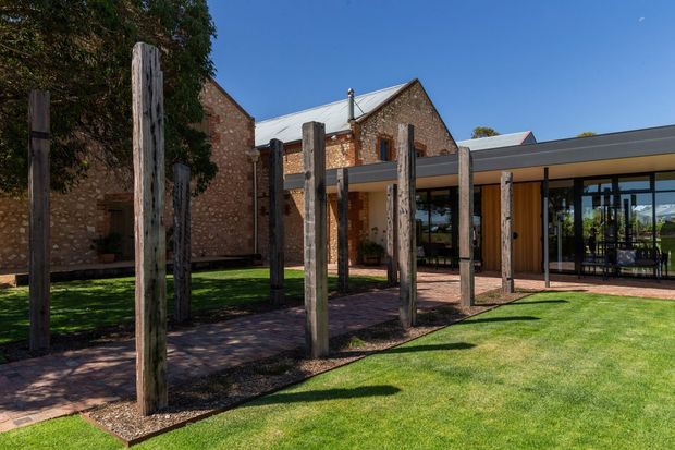 Exterior view of a renovated or extended brick building, combining an older gabled masonry structure with a contemporary low-profile glazed wing. A landscaped lawn and brick-paved courtyard sit in front, with rows of tall rustic timber posts forming a strong outdoor architectural feature. The modern wing has extensive dark-framed glazing and a shallow metal roof.